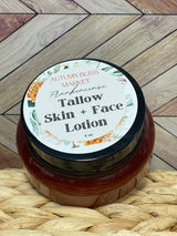 A jar of Tallow Skin + Face Lotion with a label that includes the brand name and product name, placed on a woven mat with a wooden background.