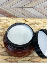 A jar of Tallow Skin + Face Lotion with a lid that shows the content of the product, placed on a woven mat with a wooden background.