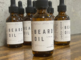 Bottles of beard oil with labels indicating 'Beard Spice' and 'Cedarwood'. Each bottle has a dropper and is labeled with the product name and the scent notes.